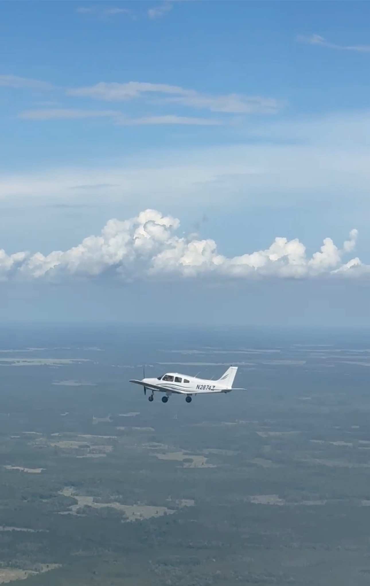 A small white propeller plane flying mid-air over a vast green landscape under a blue sky with fluffy white clouds.