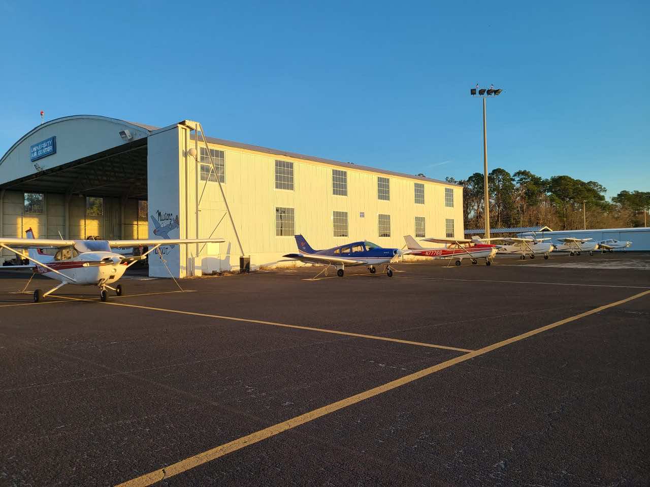 Small single-engine propeller planes parked on a tarmac in front of a white hangar under a clear blue sky.