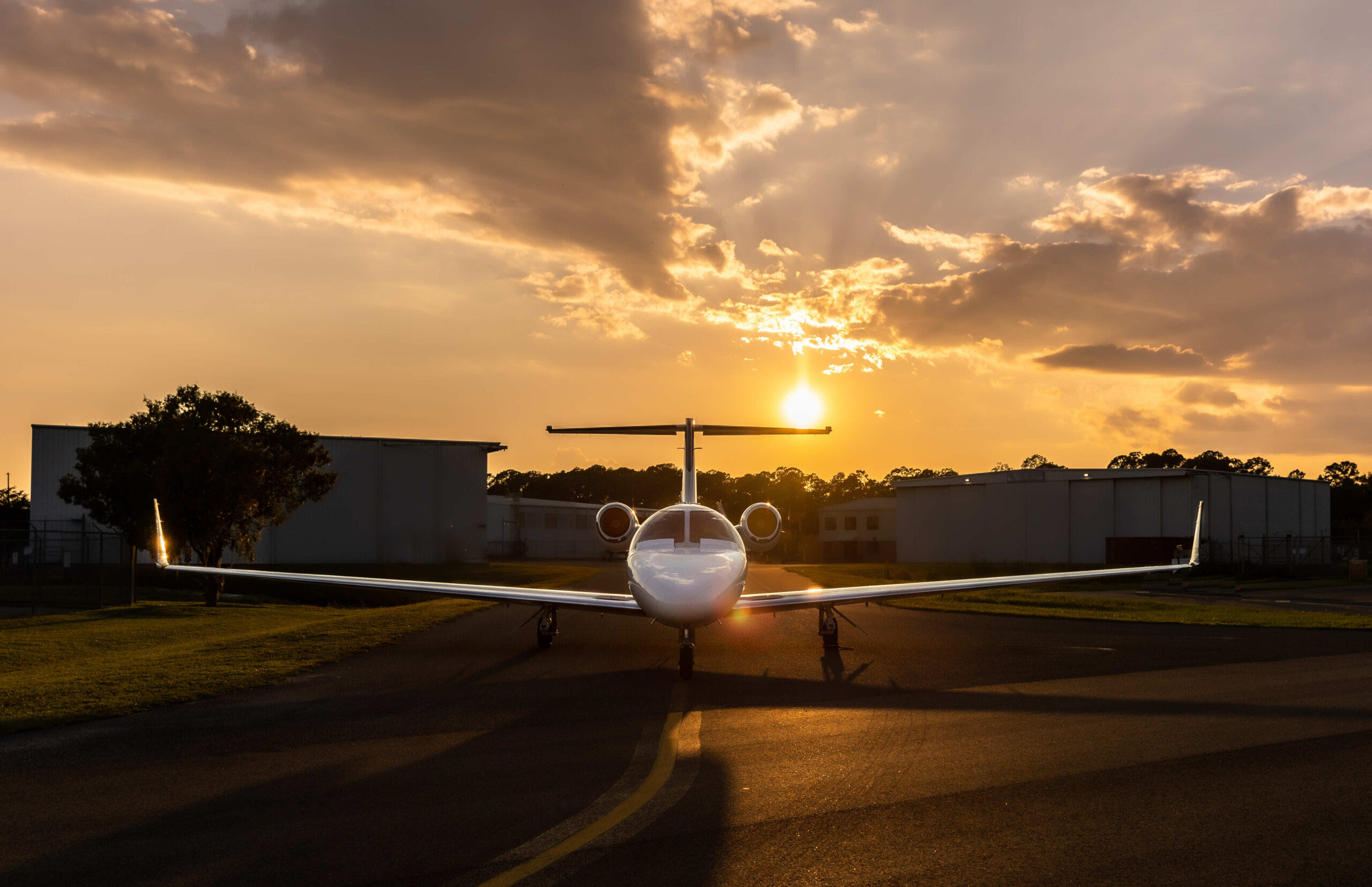 Front view of a private jet on a runway at sunset, with golden sun rays breaking through dramatic clouds.